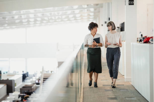 Female Tech Professionals Two female professionals having a relaxed meeting while strolling along a raised walkway in an office auditorium. human resources stock pictures, royalty-free photos & images