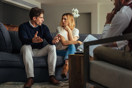 Couple arguing during therapy session with psychologist. Man and woman sitting on couch and talking while psychologist listening them.