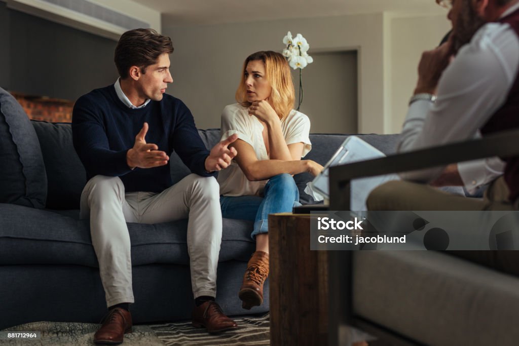 Couple arguing during therapy session with psychologist Couple arguing during therapy session with psychologist. Man and woman sitting on couch and talking while psychologist listening them. Couple - Relationship Stock Photo