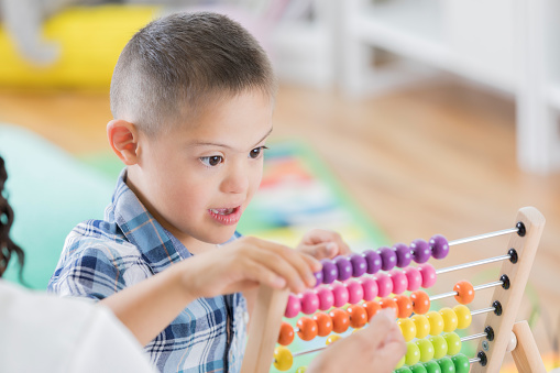 Adorable boy with special needs concentrates while counting beads on an abacus. An unrecognizable teacher is helping him.