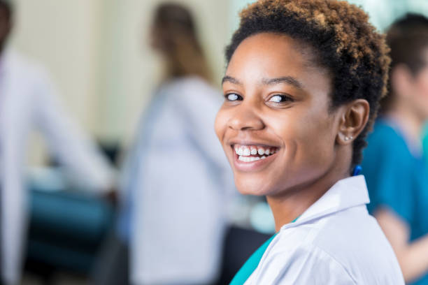 Excited young medical student smiles for camera in classroom In this closeup, a beautiful young African American medical student wears a lab coat as she turns to smile for the camera in her classroom. african american scientist stock pictures, royalty-free photos & images