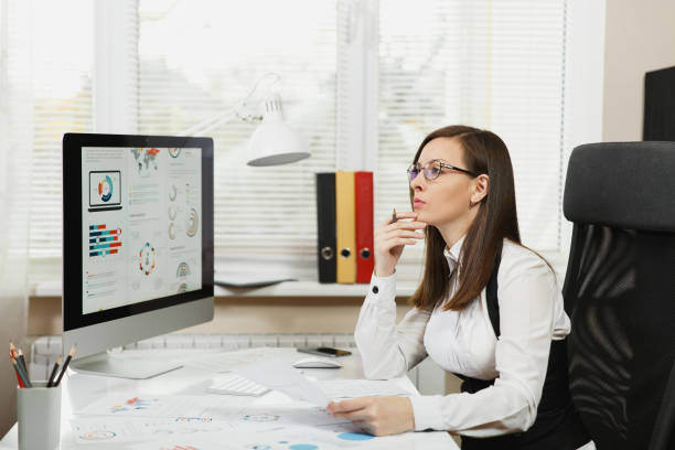 istock Beautiful business woman in suit and glasses working at computer with documents in light office, looking aside 881770100