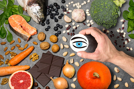Food products useful for vision. Set of natural food products are sources of vitamins and minerals. Man's hand holds tag with homemade application of eye from paper - symbol of vision. Top view