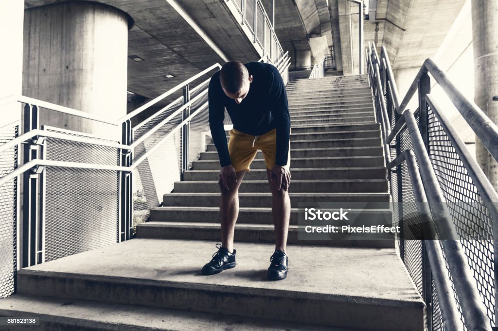 Tired male runner resting after training. Runner training and jogging outside taking a break. young handsome sports man resting after running while standing. Sport, fitness and healthy lifestyle concept. Young man exercise in urban environment. Tired Stock Photo