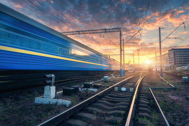 istock High speed passenger train in motion on railroad track at sunset. Railway station with blurred modern commuter train,  against colorful blue sky with red and orange clouds at dusk.Industrial landscape 881865748