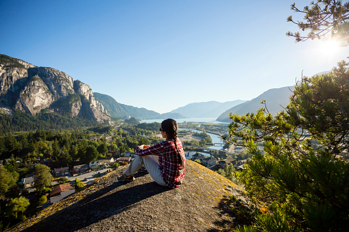 Tourist sitting on the edge of a cliff at Smoke Bluffs Park overlooking Howe Sound and the Stawamus Chief in Squamish, British Columbia, Canada.
