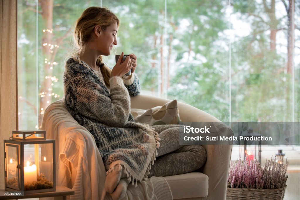 Young woman sitting home by the window Young beautiful woman sitting home in the chair by the window with cup of hot coffee wearing knitted warm sweater. Cozy room decorated with lanterns and candles. Winter Stock Photo