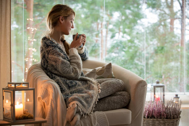 Young woman sitting home by the window Young beautiful woman sitting home in the chair by the window with cup of hot coffee wearing knitted warm sweater. Cozy room decorated with lanterns and candles. wool photos stock pictures, royalty-free photos & images