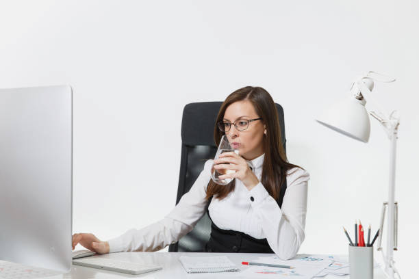 hermosa sonrisa pelo marrón mujer de negocios en traje y gafas sentado en el escritorio, beber agua pura, trabajando en equipo con moderno monitor con documentos en la oficina de luz sobre fondo blanco - office water business meeting fotografías e imágenes de stock