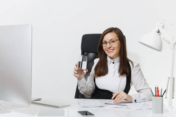 bella sonriente pelo marrón mujer de negocios en traje y gafas sentado en el escritorio con vaso de agua pura, trabajando en equipo con moderno monitor con documentos en la oficina de luz sobre fondo blanco - office water business meeting fotografías e im�ágenes de stock