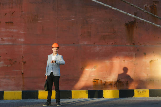 istock Handsome young unshaven successful business man in gray suit and protective construction orange helmet holding case, looking time on watch, standing in sea port against cargo rusty ship background 882186826