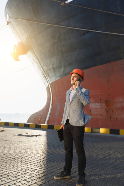 istock Handsome young unshaven successful business man in gray suit and protective construction orange helmet holding tablet, talking on mobile phone, standing in sea port against cargo rusty ship background 882187202