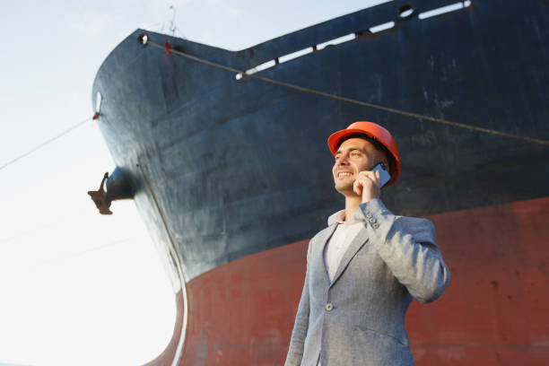 istock Handsome young unshaven successful business man in gray suit and protective construction orange helmet talking on mobile phone, standing in sea port against cargo rusty ship background. Bottom view 882187304