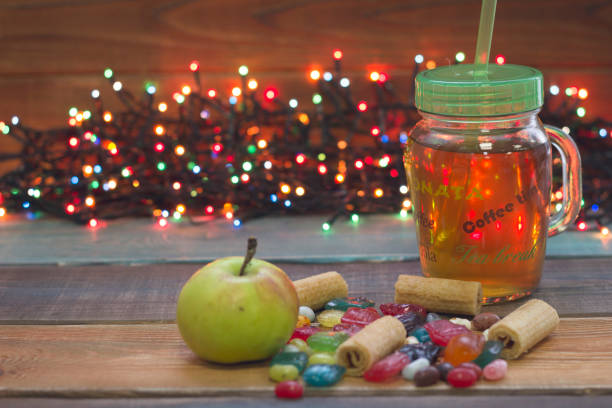 A festive still life with a glass cup of tea with a cap, green apple, candies and faity lights A festive still life with a glass cup of tea with a green plastick cap and a straw, a fresh green apple and spilled colored candies, fairy lights on the back, colored wooden background faity stock pictures, royalty-free photos & images