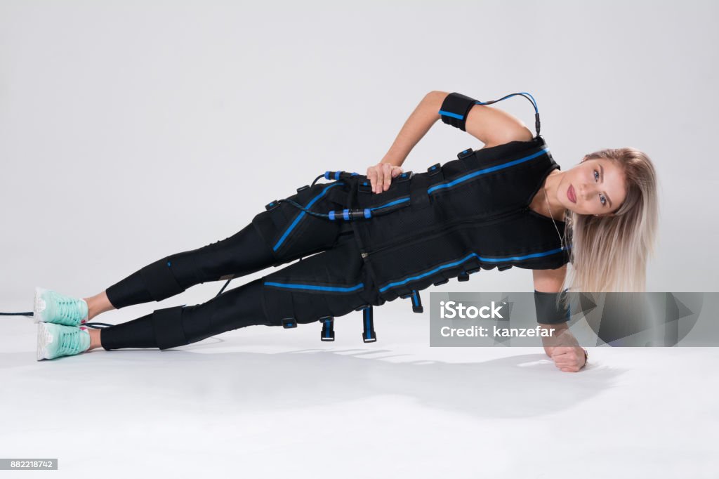 Beautiful blonde in an electric muscular suit for stimulation makes an exercise on the rug Beautiful blonde in an electric muscular suit for stimulation makes an exercise on the rug. Young woman in EMC suit isolated on white background Emergency Services Occupation Stock Photo