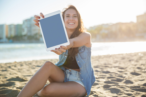 Portrait of young happy woman sitting on beach showing tablet