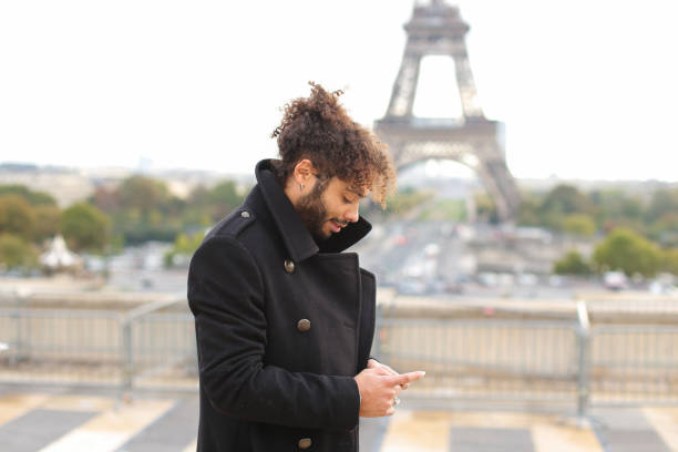 Half Italian and Nigerian man making call with modern smartphone close to Eiffel Tower in slow motion. Young male person has black curly hair, nice smile and earing, wears black coat. Concept of new technologies for young businessman, apps for talking by Internet always in touch.