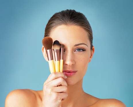 Studio portrait of a beautiful young woman holding makeup brushes in front of her eyes against a blue background
