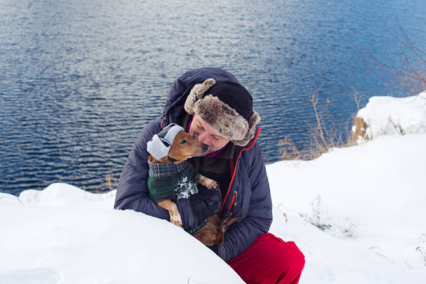 Man hugs small dog, wearing in a funny winter hat, while walking along the shore of the lake on a winter day.