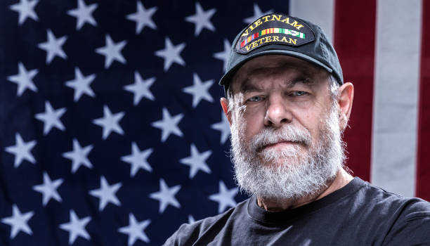 United States Navy Authentic Vietnam War Military Veteran Close-up headshot of an authentic 67 year old United States Navy Vietnam War military veteran looking at the camera. Plenty of copy space on the USA flag stars and stripes red, white and blue background. He is wearing an inexpensive, non-branded, generic, souvenir shop replica Vietnam veteran commemorative baseball hat style cap. vietnam photos stock pictures, royalty-free photos & images