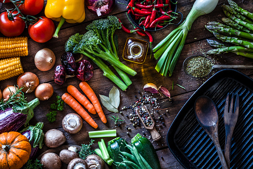 Top view of a rustic wooden table filled with fresh organic vegetables ready for cooking. At the bottom-right is a cast iron pan with a fork and spoon inside. The composition includes garlic, tomatoes, asparagus, edible mushrooms, eggplant, cucumber, broccoli, onion, carrot, corn, and chili peppers. DSRL studio photo taken with Canon EOS 5D Mk II and Canon EF 100mm f/2.8L Macro IS USM