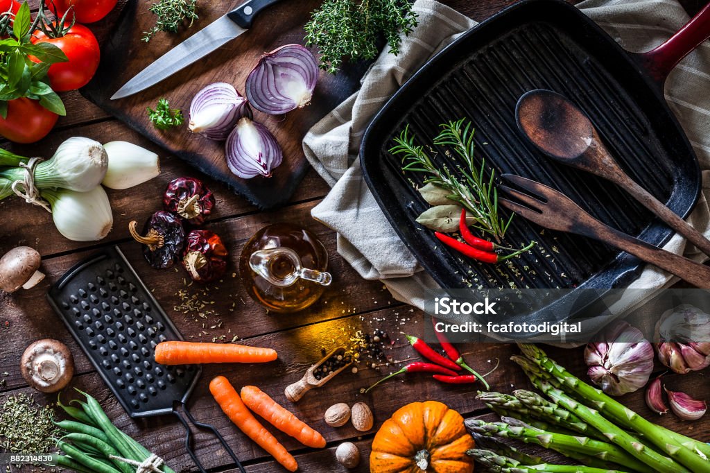 Fresh vegetables ready for cooking shot on rustic wooden table Top view of a rustic wooden table filled with fresh organic vegetables ready for cooking. At the top-right is a cast iron pan and beside it is a cutting board with some spanish onions. The composition includes garlic, tomatoes, asparagus, edible mushrooms, eggplant, cucumber, broccoli, onion, carrot, corn, and chili peppers. DSRL studio photo taken with Canon EOS 5D Mk II and Canon EF 100mm f/2.8L Macro IS USM Cooking Stock Photo