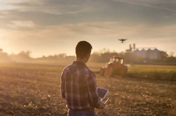 Attractive farmer navigating drone above farmland with silos and tractor in background. High technology innovations for increasing productivity in agriculture