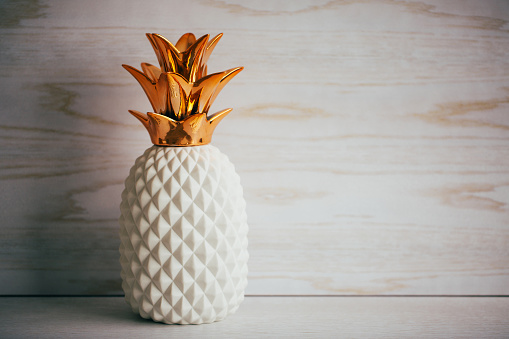 Decorative ceramic pineapple with golden leaves on white wooden background.