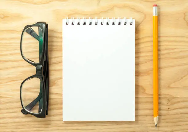 Top view of open spiral blank notebook, glasses and pencil on wood desk background