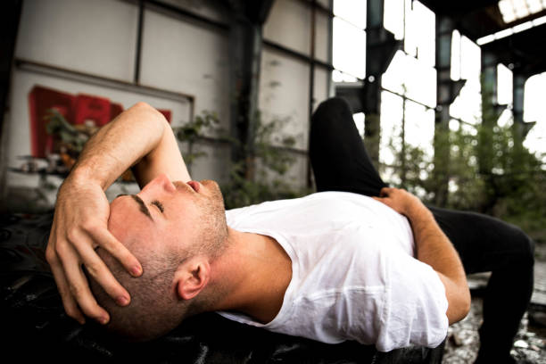 Young man in white t-shirt and black trousers lying outdoors, holding his head with hand and looking up with sad facial expression.