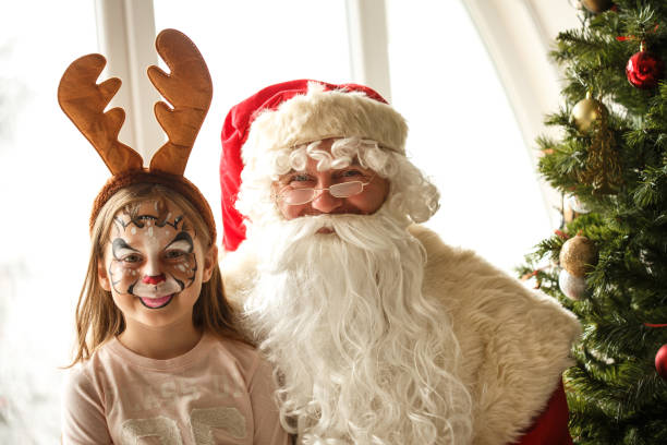 Cute, little girl with Reindeer  face paint and Santa Claus sitting next to a Christmas tree.