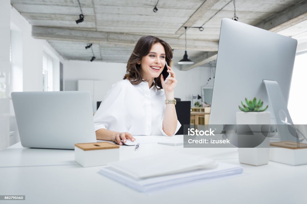 Businesswoman working in modern workplace Young businesswoman sitting at her desk talking on phone and working on computer. Female executive working in modern workplace. Office Stock Photo