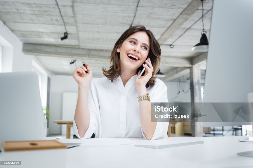 Smiling businesswoman making a phone call in office Cheerful young beautiful woman talking on mobile phone while sitting at her working place. Smiling businesswoman making a phone call in office. Adult Stock Photo