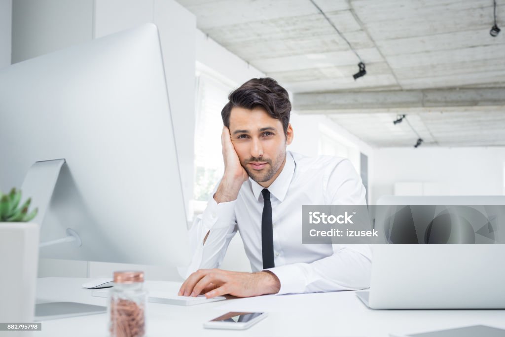 Handsome young businessman sitting at his desk Handsome young businessman sitting at his desk and looking at camera. Caucasian male executive at work in modern workplace. Boredom Stock Photo
