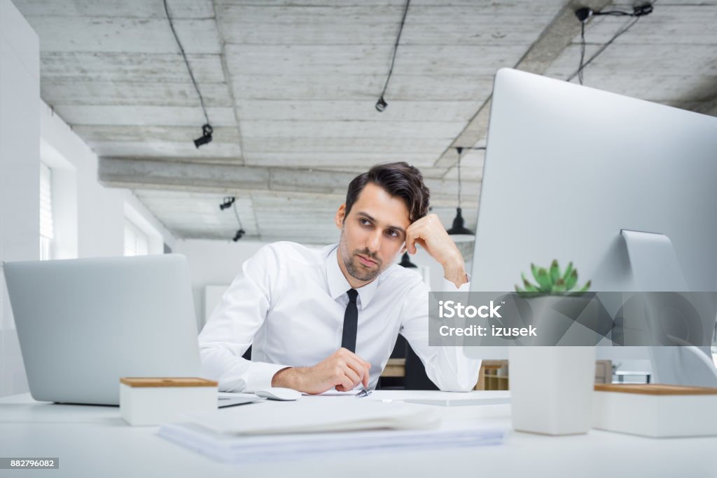 Pensive businessman sitting at his office desk Pensive businessman sitting at his office desk. Worried male business professional sitting at table and looking away in thought. Adult Stock Photo