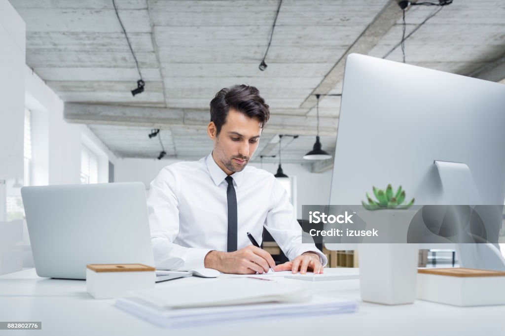 Young businessman working in office Young caucasian businessman working in office. Male executive sitting at his desk and making notes. Adult Stock Photo
