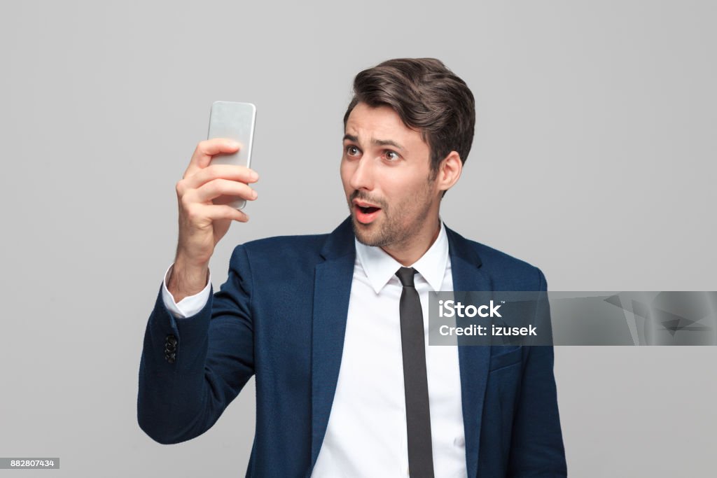 Surprised businessman looking at mobile phone Surprised young businessman in suit looking at mobile phone while standing against grey background 25-29 Years Stock Photo