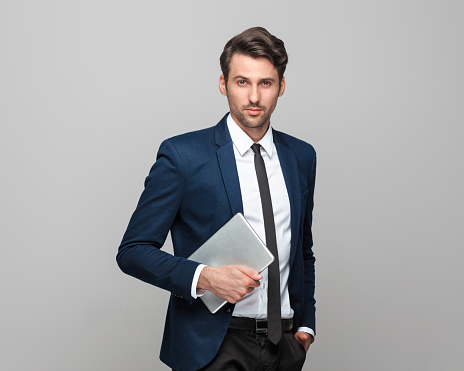 Portrait of handsome businessman holding digital tablet standing against grey background and looking at camera