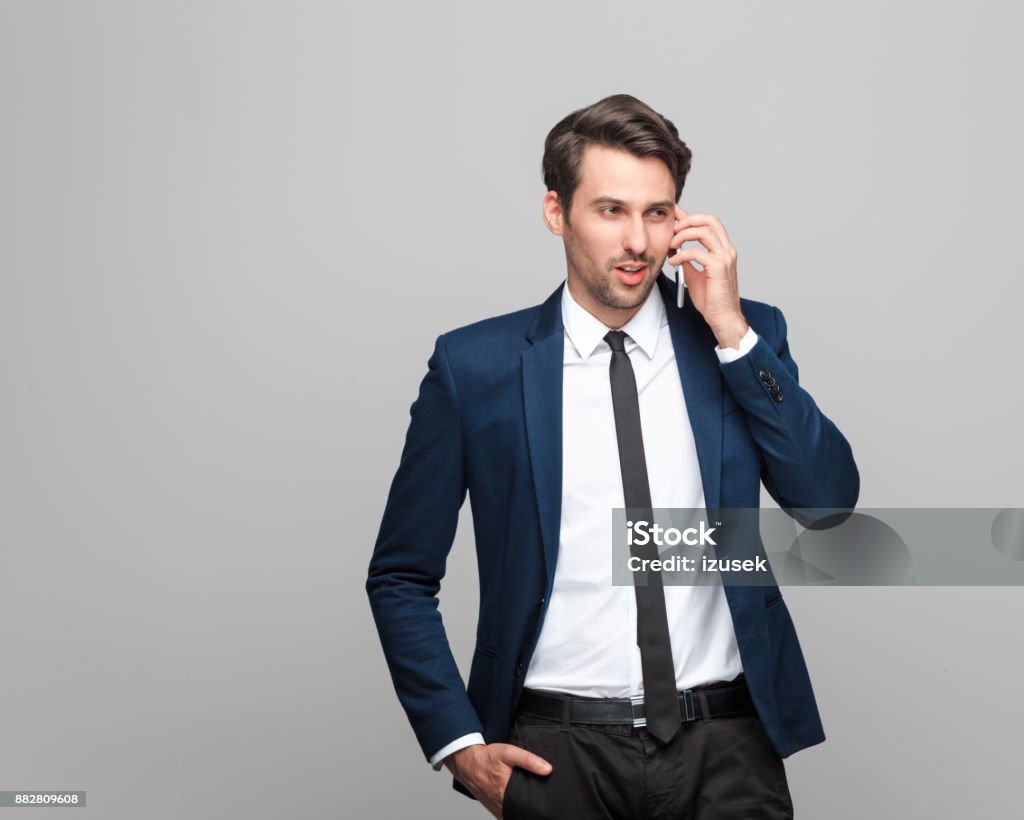 Young businessman talking on mobile phone Portrait of young businessman talking on mobile phone while standing on grey background Business Stock Photo