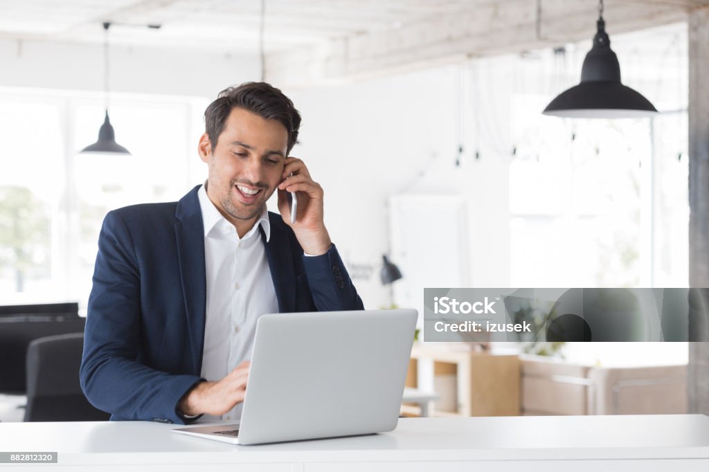 Businessman working at his desk Happy young businessman talking on phone and using laptop in office. Caucasian male executive working at his desk. Suit Stock Photo