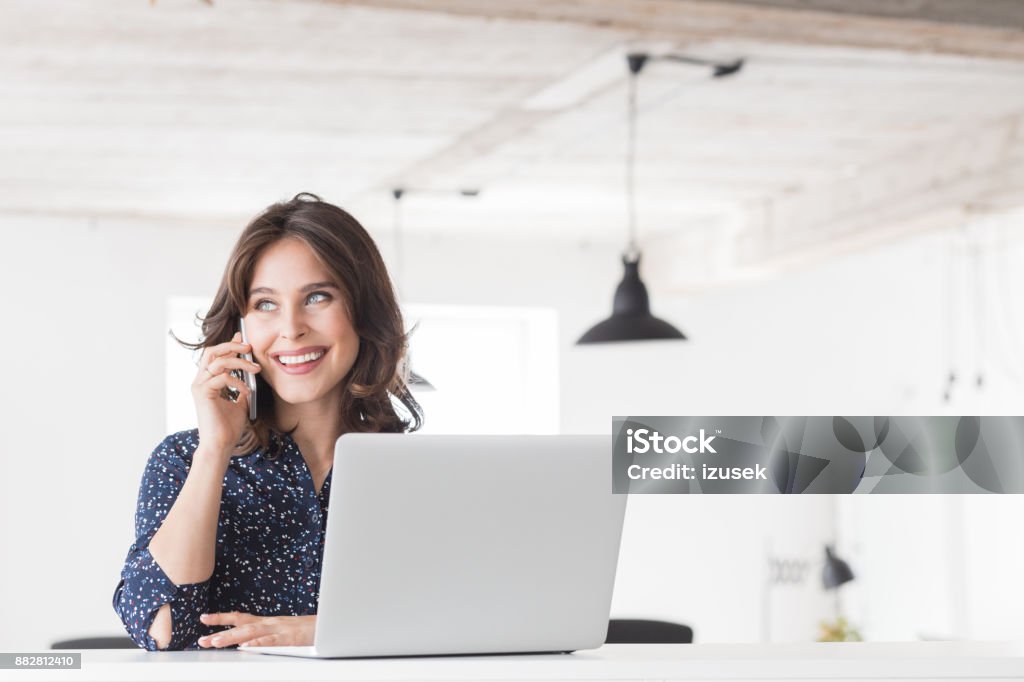 Beautiful woman talking on phone at office Beautiful young woman sitting at her desk with laptop and talking on mobile phone. Female working at her office desk making a phone call. Businesswoman Stock Photo