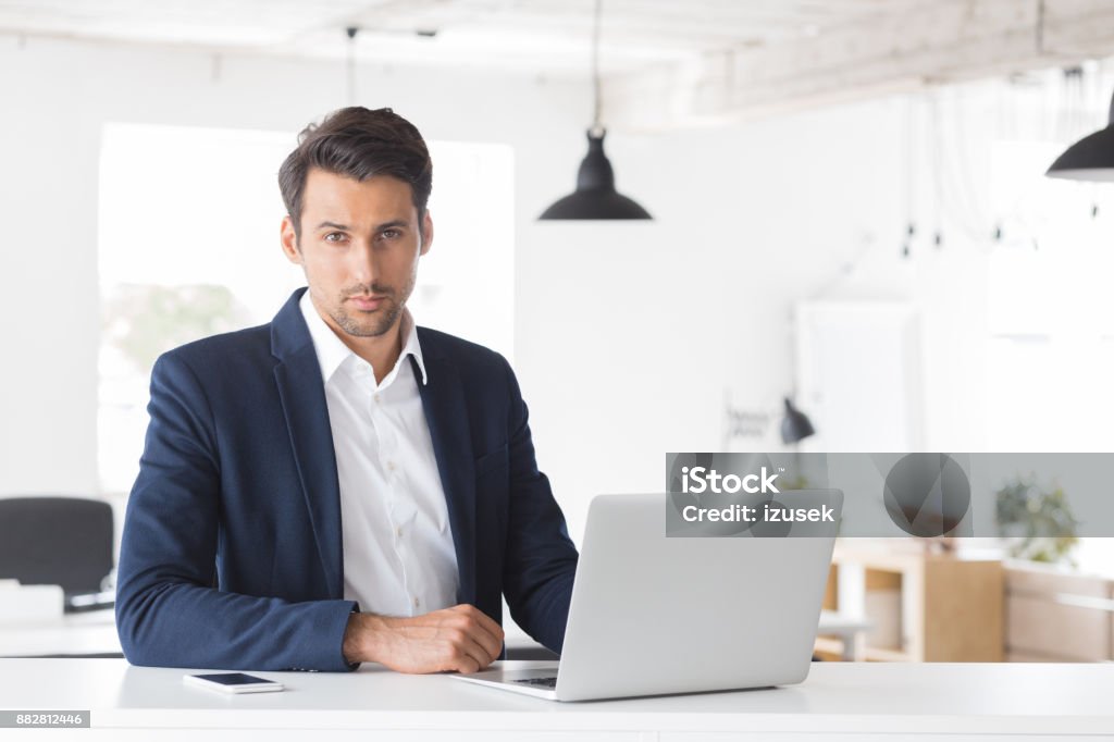 Businessman sitting at his desk with laptop Young businessman sitting at his desk with laptop. Caucasian male executive looking at camera while sitting in modern workplace. Desk Stock Photo