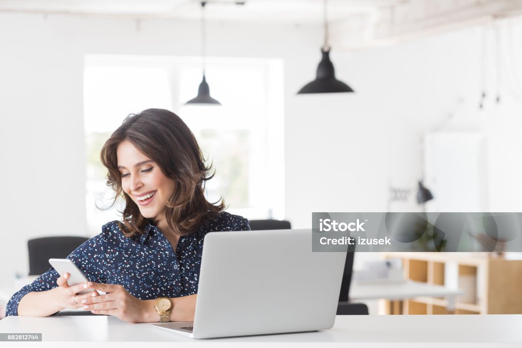 Smiling young woman using phone at her office desk Smiling young woman sitting at her desk with laptop and reading mobile phone. Female working at her office desk. 20-24 Years Stock Photo