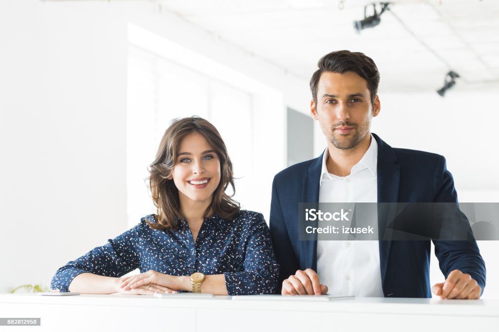 Confident business colleagues together in office Two young businesspeople standing together in office. Confident businessman and businesswoman looking at camera. 20-29 Years Stock Photo