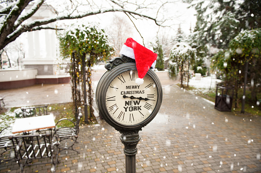 vintage street clock with the inscription Merry Christmas New York and Santa Claus hat on them outdoors in winter park with falling snowflakes
