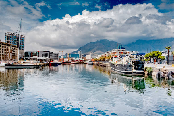 A sunny Cape Town harbor scene featuring sailboats and yachts moored in a marina, with modern waterfront apartments and the Twelve Apostles mountains in the distance.
