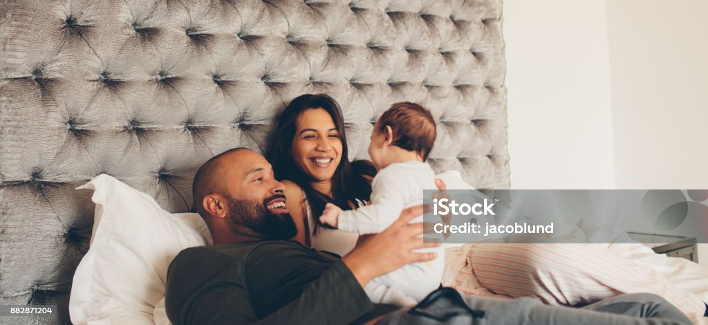 Happy parents playing with their newborn son on bed Happy parents playing with their newborn son on bed at home. Couple with little baby boy on bed. Family Stock Photo