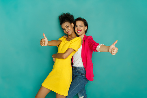 Multiethnic friendship. Happy and caucasian girls in colorful casual clothes showing thumb up at blue studio background with copy space looking at camera