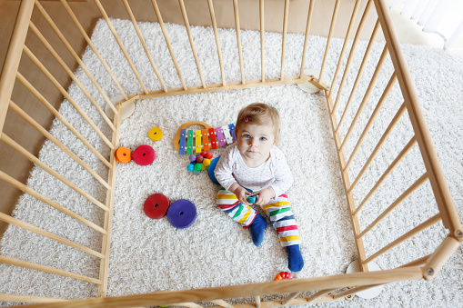 Beautiful little baby girl sitting inside playpen. Cute adorable child playing with colorful toys. Home or nursery, safety for kids.