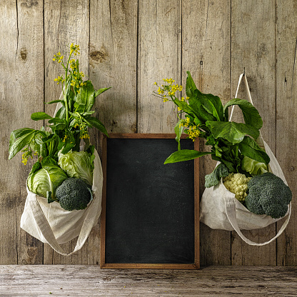Leafy green vegetables hanging in reusable cotton bags, either side of a blackboard, which is between the two bags, set on an old wood board wall background. Vegetables include choy sum, cauliflower, broccoli and Chinese cabbage.